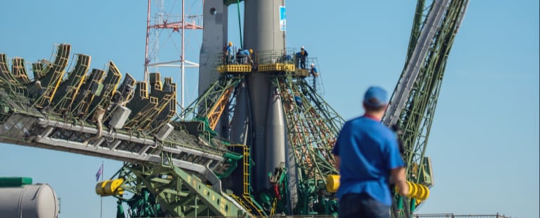 A rocket on a launch pad with a person in a blue shirt and cap standing in the foreground, set against a clear blue sky.